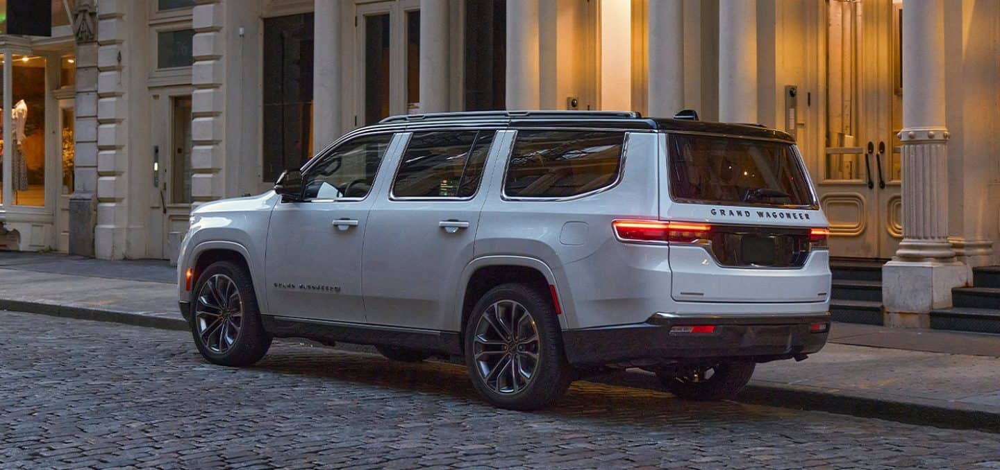 Display The 2023 Grand Wagoneer Series III with its taillamps lit as it is stopped at the curb on a cobble-stoned street.