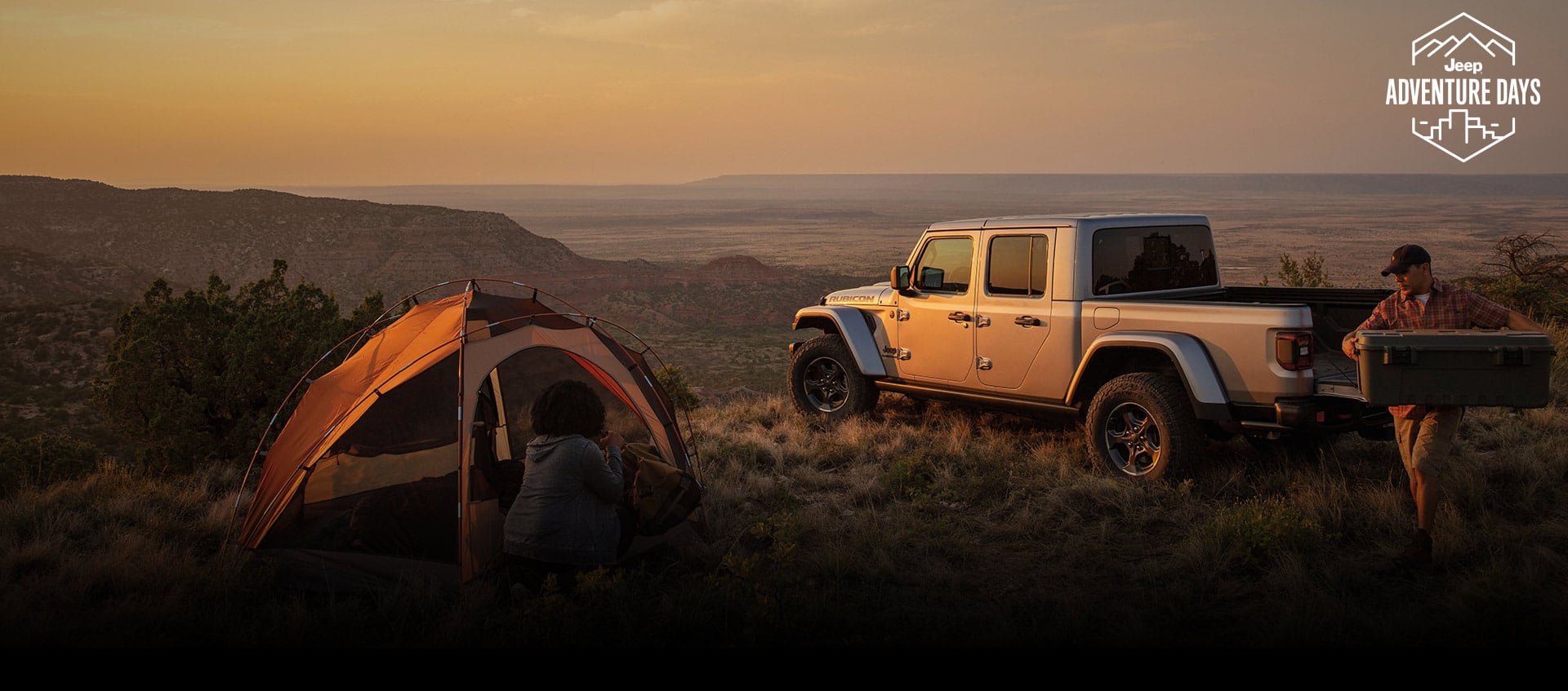A driver-side rear profile of a 2023 Jeep Gladiator Rubicon parked on a hilltop at dusk with its tailgate open and a man carrying a cooler to a nearby campsite. Jeep Adventure Days logo.