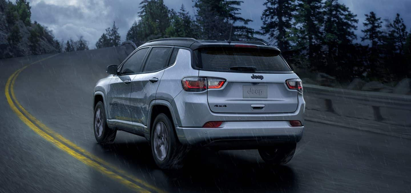 Display A rear view of a 2024 Jeep Compass High Altitude being driven on a winding highway during a rainstorm.