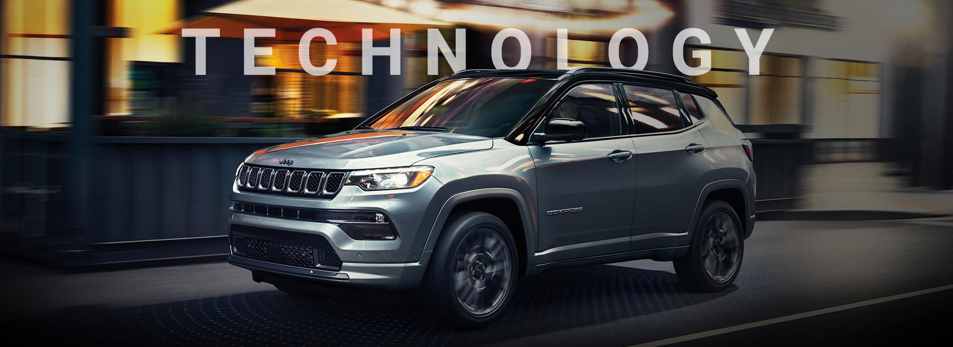 A silver 2024 Jeep Compass High Altitude being driven on a city street at night with the background blurred to indicate the vehicle is in motion. Technology.