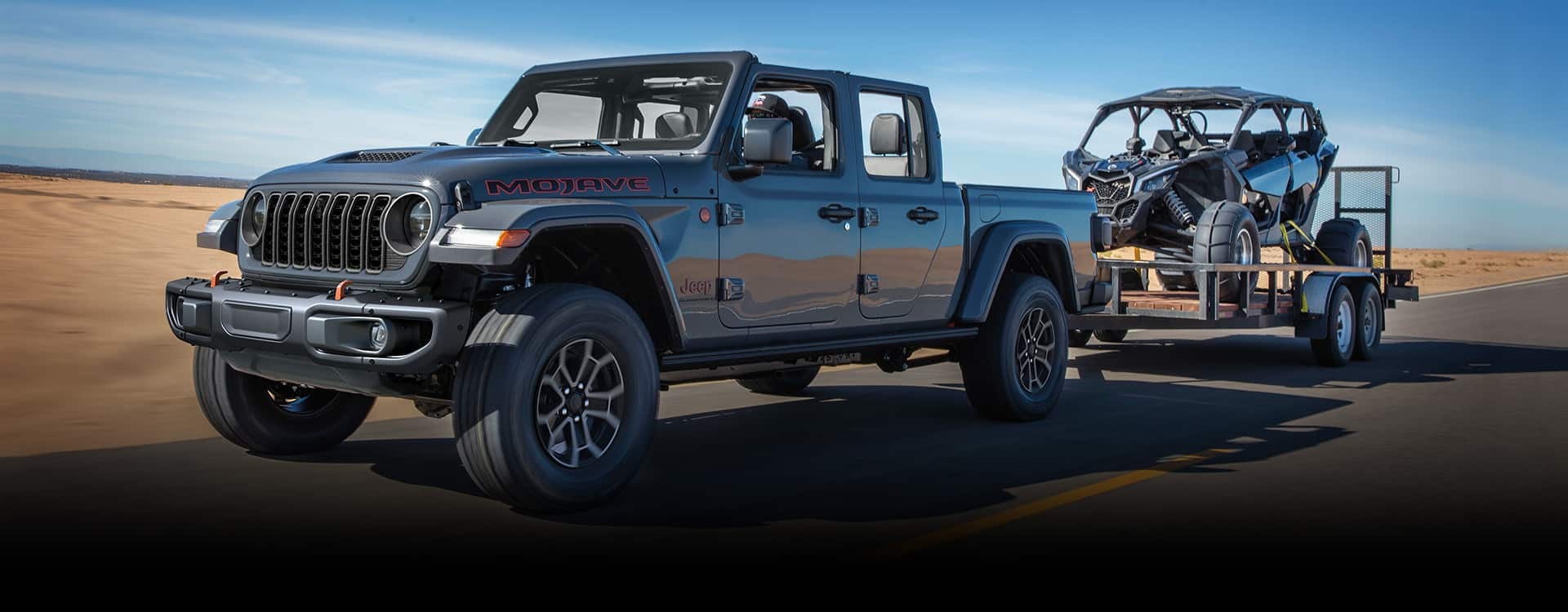 A gray 2024 Jeep Gladiator Mojave being driven down a highway as it tows an ATV on a utility trailer.