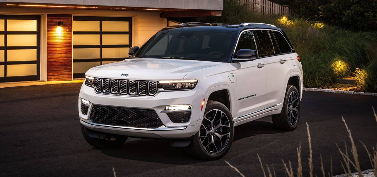 Display A white 2024 Jeep Grand Cherokee Summit Reserve 4xe parked outside of a contemporary home in the evening. 