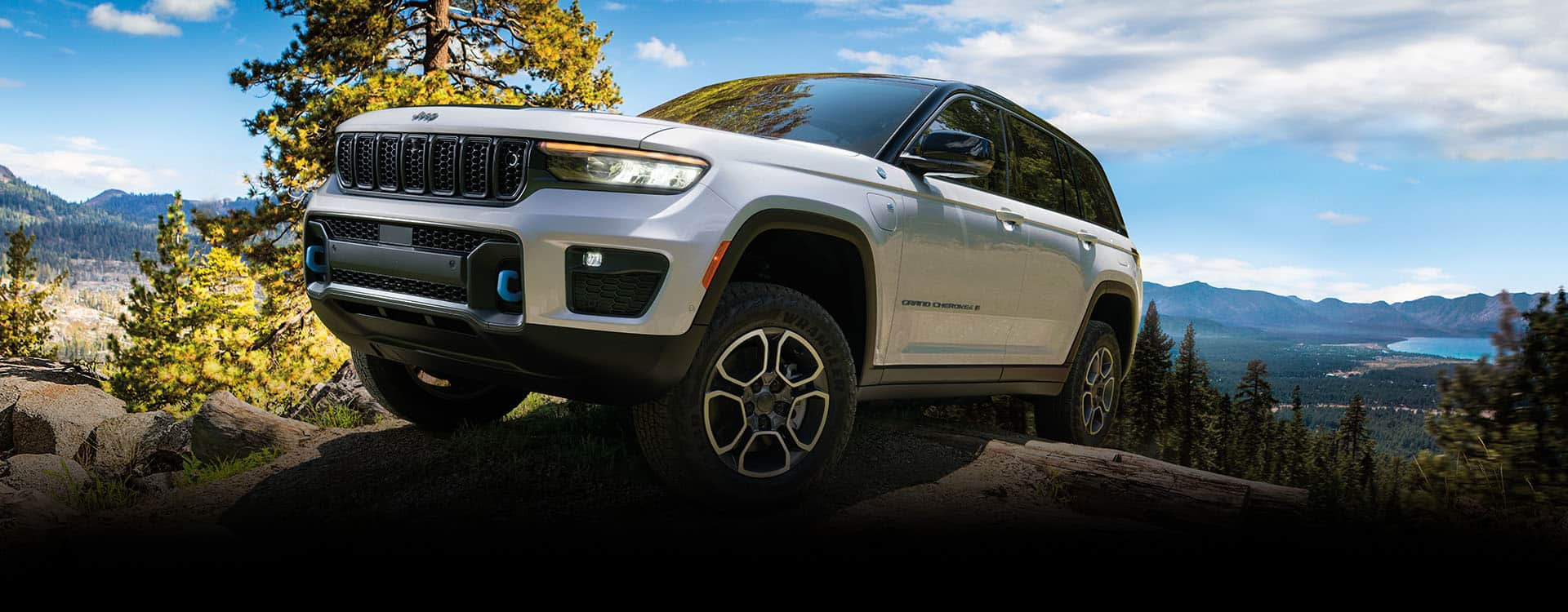 A white 2024 Jeep Grand Cherokee Trailhawk 4xe with black hood insert, being driven over an outcropping, with mountains and a lake in the background.